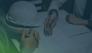 Three people reviewing technical engineering drawings on a table, one holding a pen near a hard hat, discussing CMMC scope and compliance