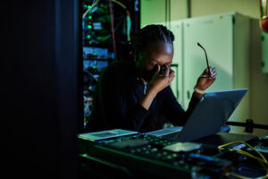 A frustrated cybersecurity expert pinches the bridge of their nose as they work in a server room.