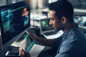 young man at a desk using a computer and tablet to assess and manage a company’s cybersecurity.