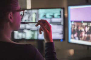 View over the shoulder of a young woman looking at two computer screens, managing her business' cybersecurity.